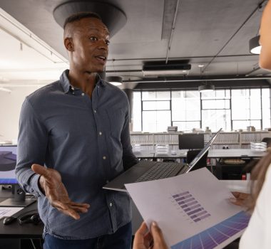 Diverse coworkers discussing printed bar-chart reports at desk in open plan office with laptop
